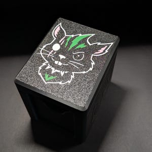 Può includere: Scatola nera con una parte superiore strutturata e glitterata con un design a gatto dei cartoni animati. Il gatto ha accenti verdi e bianchi, con un orecchio rosa e un contorno bianco. La scatola è angolata.