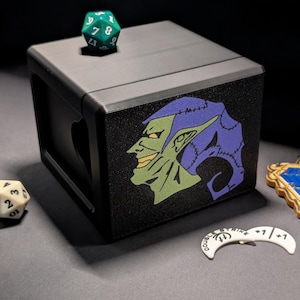 May include: A black dice box featuring a green and purple goblin design, a green 8-sided die, a white 20-sided die, a gold and blue charm, and a white game token. The dice box has a heart-shaped cutout.