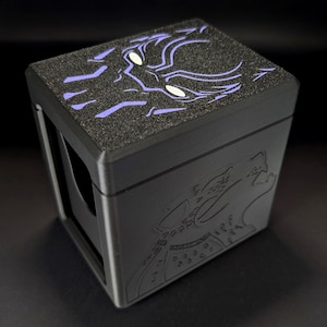 Puede incluir: Una caja de almacenamiento rectangular negra con una tapa texturizada que presenta un gráfico morado y blanco de una máscara de pantera estilizada. El frente de la caja muestra el contorno en relieve de una cabeza de pantera y el lateral tiene una abertura.