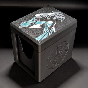 Peut inclure: Une boîte de rangement rectangulaire noir mat avec une surface supérieure scintillante ornée d'une illustration graphique stylisée bleue et blanche. Le côté de la boîte présente un emblème circulaire en relief avec un motif de soleil.