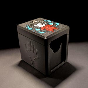 Könnte beinhalten: Schwarze Kartenaufbewahrungsbox mit einer farbenfrohen Illustration auf der Oberseite. Die Illustration zeigt eine Cartoonfigur in Rot und Blau. Die Box hat einen Schlitz auf der Vorderseite und ein Symbol an der Seite. Die Box ist für Sammelkartenspiele konzipiert.