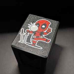Könnte beinhalten: Schwarze Box mit einer strukturierten, glitzernden Oberseite mit einer Cartoon-Figur von Deadpool, die eine Blase bläst und zwei Schwerter hält. Die Figur ist rot und schwarz mit weißen Akzenten. Die Box steht auf einer dunklen Oberfläche.