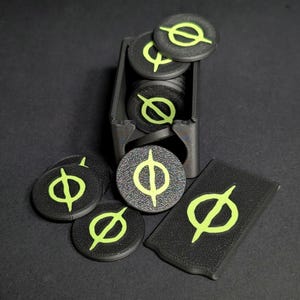 Könnte beinhalten: Ein schwarzer rechteckiger Aufbewahrungsbehälter gefüllt mit runden schwarzen Spielsteinen, die ein neongrünes Phi-Symbol tragen. Mehrere zusätzliche runde Spielsteine und ein rechteckiger Spielstein mit demselben neongrünen Symbol liegen davor.