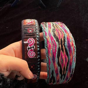 May include: Two colorful embroidered bands, one with a geometric pattern and the other with a floral design. The bands are made of fabric and have a black background.