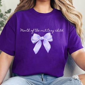 May include: A purple t-shirt with the text "Month of the military child" and a light purple bow graphic. The shirt is a short-sleeved crew neck style.