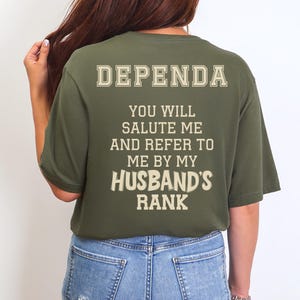 May include: Olive green t-shirt with the word "DEPENDA" printed across the upper back in a vintage style font. Below, text reads "YOU WILL SALUTE ME AND REFER TO ME BY MY HUSBAND'S RANK". The shirt is worn by a person wearing blue jeans.