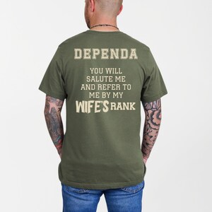 A man wearing a green t-shirt with large cream-colored text on the back reading: “DEPENDA. YOU WILL SALUTE ME AND REFER TO ME BY MY WIFE’S RANK.” The text is bold and blocky, arranged in a playful, military-inspired style.