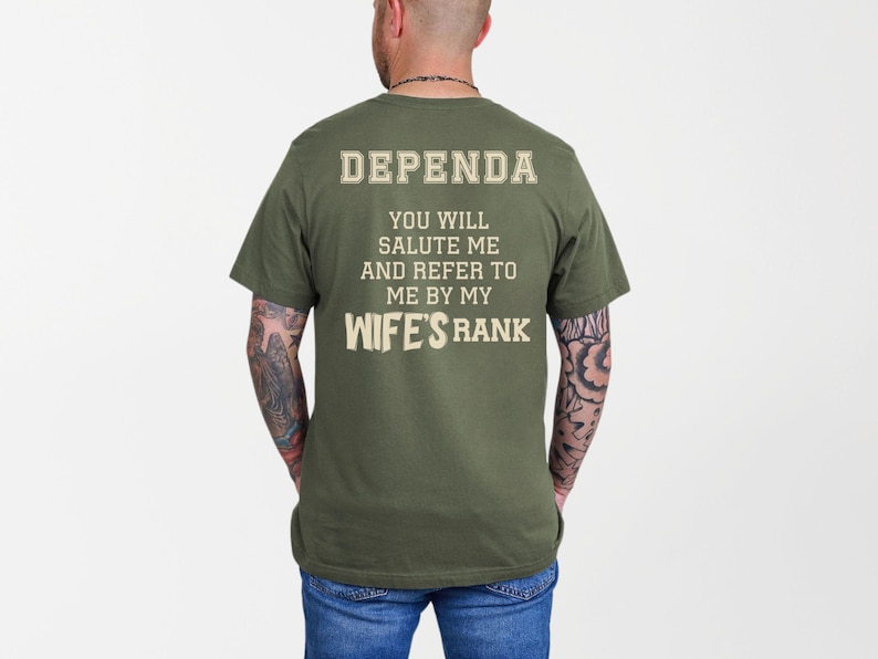 A man wearing a green t-shirt with large cream-colored text on the back reading: “DEPENDA. YOU WILL SALUTE ME AND REFER TO ME BY MY WIFE’S RANK.” The text is bold and blocky, arranged in a playful, military-inspired style.