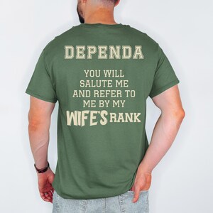 A man wearing a green t-shirt with large cream-colored text on the back reading: “DEPENDA. YOU WILL SALUTE ME AND REFER TO ME BY MY WIFE’S RANK.” The text is bold and blocky, arranged in a playful, military-inspired style.