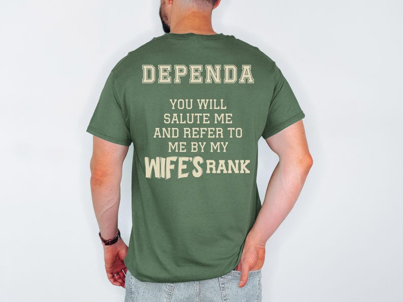 A man wearing a green t-shirt with large cream-colored text on the back reading: “DEPENDA. YOU WILL SALUTE ME AND REFER TO ME BY MY WIFE’S RANK.” The text is bold and blocky, arranged in a playful, military-inspired style.