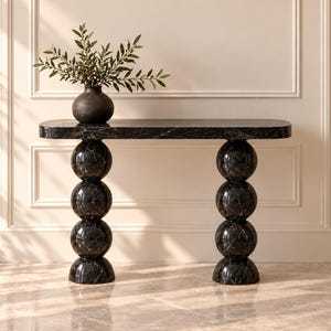 May include: A black marble console table with a smooth rectangular top and two legs composed of stacked marble spheres. A dark, rounded vase containing green leafy branches sits on the left side of the table surface.