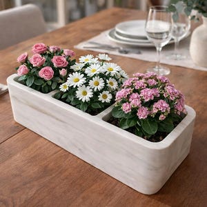 Puede incluir: Una jardinera rectangular con efecto mármol que contiene tres tipos de plantas con flores. Rosas rosas, margaritas blancas y kalanchoes rosas están dispuestas en compartimentos separados. La jardinera está sobre una mesa de madera, con vajilla en el fondo.