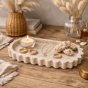 Op de afbeelding: Een beige stenen dienblad met een geschulpte rand bevat gouden sieraden, een brandende kaars en decoratieve items. De sieraden omvatten twee oorringen, een hangerketting en een ring. Droogbloemen en een vaas staan op de achtergrond.