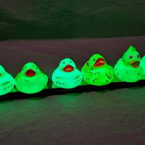 May include: A row of 10 glowing rubber duckies in various colors and designs, including a red duck, a green duck with a cat face, and a green duck with a skull and crossbones design.