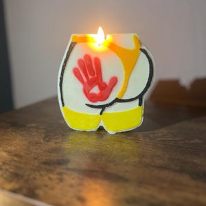 May include: A yellow and white candle shaped like a human bottom with a red handprint on the back. The candle is lit and the flame is visible.
