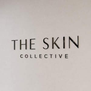 May include: A wall-mounted sign featuring the black, three-dimensional text THE SKIN COLLECTIVE in a clean, sans-serif font. The letters are mounted on a smooth, light-coloured wall, creating a professional and clear display for the brand name.