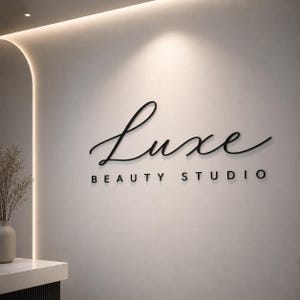 May include: A wall-mounted sign featuring the word Luxe in a black cursive script font, positioned above the words BEAUTY STUDIO in a black sans-serif font. The lettering is three-dimensional and mounted on a smooth, light-coloured wall surface.