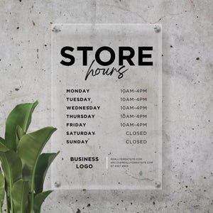 Op de afbeelding: Een helder acrylbord met openingstijden, bevestigd aan een grijze, getextureerde muur. Het bord leest "STORE hours" in zwart, met de dagen van de week en openingstijden. Een logo en contactgegevens zijn ook opgenomen.