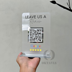 May include: A clear acrylic sign with the text "LEAVE US A Review and receive £5 off of your next visit!"  A black and white QR code is displayed below the text. The sign also features the Google logo and five yellow stars.