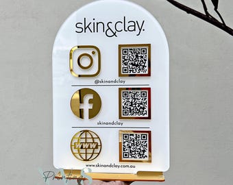 Op de afbeelding: Een wit acryl bord met gouden letters en pictogrammen. Het bord staat "skin&clay". Het bord bevat pictogrammen voor Instagram, Facebook en een website, samen met QR-codes voor elk.