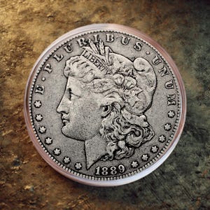 Puede incluir: Una moneda de plata con el perfil de una mujer, con las palabras "LIBERTY" y "E PLURIBUS UNUM" visibles. La moneda está fechada en 1889 y está encapsulada en un soporte de plástico transparente. El diseño es detallado, sobre un fondo texturizado.