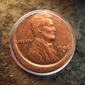 Puede incluir: Una moneda de cobre de 1946 de Lincoln en un soporte de plástico transparente. La moneda presenta un perfil de un hombre y la palabra "LIBERTY" encima del año. La moneda está sobre un fondo marrón texturizado.