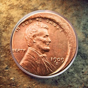 Puede incluir: Un centavo Lincoln de 1909 de color cobre en un soporte transparente. La moneda muestra un perfil de Abraham Lincoln y las inscripciones "IN GOD WE TRUST" y "LIBERTY". El año "1909" es visible.