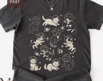 May include: A charcoal gray crew neck t-shirt featuring a cream-colored graphic print of multiple cats wearing glass space helmets, surrounded by small stars and planets. The design is centered on the front of the shirt, which has short sleeves.