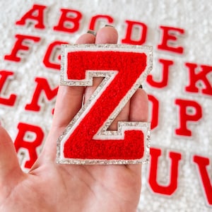 May include: Red felt letter Z with a white outline and silver glitter trim. The letter is held in a hand against a background of other letters of the alphabet.