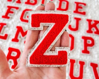 May include: Red felt letter Z with a white outline and silver glitter trim. The letter is held in a hand against a background of other letters of the alphabet.
