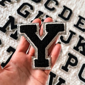 May include: A black felt letter Y with a silver glitter outline. The letter is held in a hand against a background of other letters.