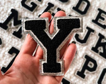 May include: A black felt letter Y with a silver glitter outline. The letter is held in a hand against a background of other letters.