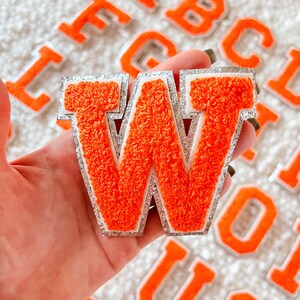 May include: Orange felt letter W with a silver glitter trim. The letter is held in a hand against a background of other felt letters.