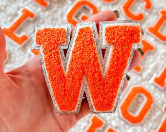 May include: Orange felt letter W with a silver glitter trim. The letter is held in a hand against a background of other felt letters.