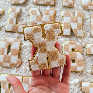 May include: A large, fluffy, tan and white checkered letter X with a gold outline. The letter is held in a hand against a white fuzzy background.