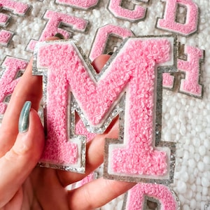 May include: Pink felt letter M with silver glitter trim. The letter is surrounded by other letters in the background.