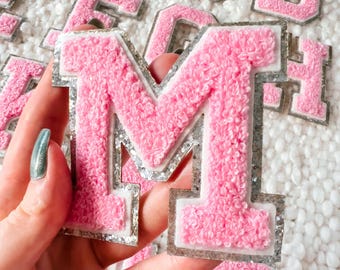 May include: Pink felt letter M with silver glitter trim. The letter is surrounded by other letters in the background.