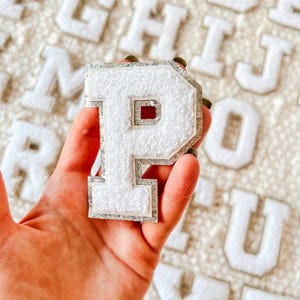 May include: White felt letter P with a silver outline. The letter is held in a hand against a background of other white felt letters.
