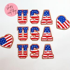 May include: Patches with the letters "USA" in red, white, and blue, resembling the American flag. The letters have a gold glitter border. Two heart-shaped patches with the American flag design are also included.