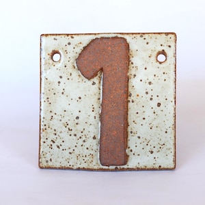 May include: A square ceramic house number tile featuring the number 1 in a dark brown, textured clay finish. The background of the tile is a glossy, off-white glaze with brown speckles throughout. Two circular holes are positioned at the top corners.