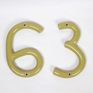 May include: Two glossy, olive-green ceramic house numbers, specifically the digits 6 and 3. Each number features a smooth, rounded surface with two small, circular mounting holes positioned at the top and bottom for secure installation.