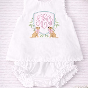 Peut inclure: Ensemble de vêtements blancs pour bébé, composé d'un haut sans manches et d'un bloomer à volants. Le haut présente un écusson brodé avec les initiales "SEG" en rose, flanqué de deux illustrations de chiens avec des nœuds roses. La marque "Kenton Co Handmade" est en bas.