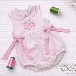 May include: A light pink seersucker romper with a Peter Pan collar and embroidered initials. The baby garment features pink bows at the waist and is displayed with spools of pink and green thread. The romper is handmade.