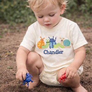 Puede incluir: Un niño pequeño con un body color crema y una camiseta de manga corta con insectos bordados y el nombre "Chandler". El niño está jugando con insectos de juguete de colores. La camiseta presenta insectos naranjas, verdes, azules y turquesas.