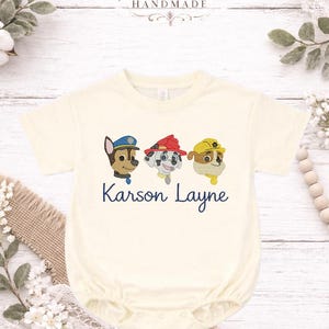 May include: A cream-coloured baby onesie featuring embroidered cartoon dog characters wearing hats. The text "Karson Layne" is embroidered in navy blue below the characters. The top of the image has the text "Kenton Co Handmade".