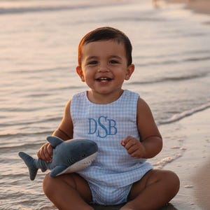May include: A smiling child wearing a light blue gingham romper with the embroidered initials "DSB" and holding a gray shark plush toy. The child is sitting on a sandy beach near the water's edge.