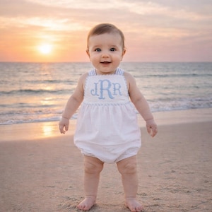 Puede incluir: Un bebé con un pelele blanco con iniciales bordadas en azul está de pie en una playa de arena al atardecer. El pelele tiene una tela texturizada y ribetes a rayas azules. El bebé sonríe, con el mar al fondo.