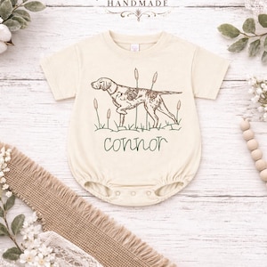 May include: Cream-colored baby onesie with a brown dog illustration, cattails, and the name "Connor" in a script font. The top of the onesie has the text "Kenton Co Handmade" in a decorative font. The onesie is on a white wooden background.