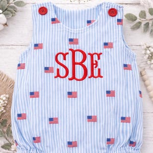 May include: Blue and white striped baby romper with red embroidered initials and American flag accents. Features red buttons at the shoulders. The brand name "Kenton Co Handmade" is visible at the top.