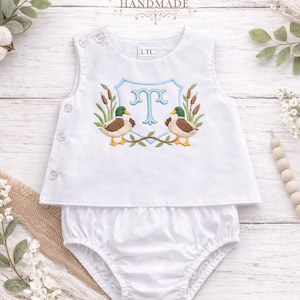 May include: White baby outfit with a top and diaper cover. The top features an embroidered design with two ducks, cattails, and a blue monogram. The brand name "Kenton Co Handmade" is at the top.